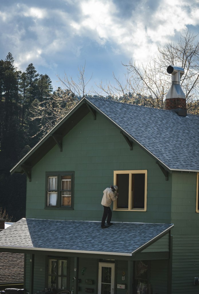 Painter working on window trim of a green Craftsman house, illustrating exterior painting in Oregon